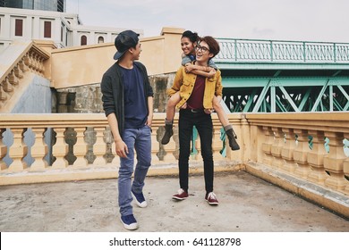 Joyful Asian teenager piggybacking his pretty girlfriend while walking with their friend in city and chatting animatedly
