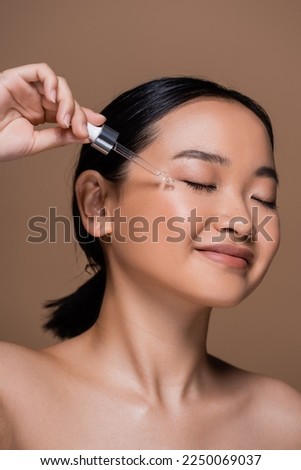 Joyful asian woman with dropper applying cosmetic serum on cheek isolated on brown