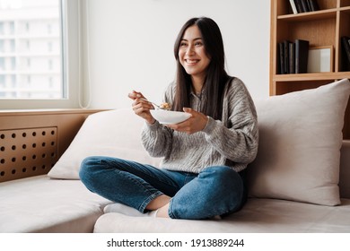 Joyful Asian Woman Smiling And Eating While Sitting On Couch At Home