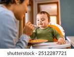 Joyful baby boy in green outfit being spoon-fed by loving mother. Adorable toddler enjoying a healthy meal in high chair at home.