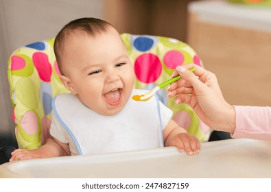 A joyful baby boy sits in a colorful high chair, eagerly opening their mouth as a caregiver feeds them with a spoon of baby food in a bright, cheerful kitchen. - Powered by Shutterstock