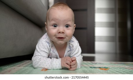 Joyful baby crawls on the mat and smiles. A child is crawling on his stomach. Baby boy playing with toy on the floor. Smiling baby lies on his stomach and looking at camera on the play mat. 