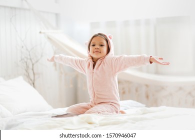 Joyful baby girl in warm pink pajamas with ears on the bed. A child in plush pajamas on a light bedroom background, the concept of a good cozy children's morning