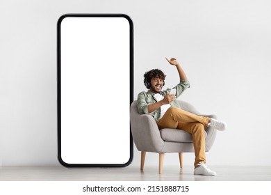 Joyful Bearded Curly Indian Guy In Casual Sitting In Armchair With Brand New Smartphone And Wireless Headset, Watching Movie Or Listening To Music Online While Chilling At Home, Copy Space, Mockup