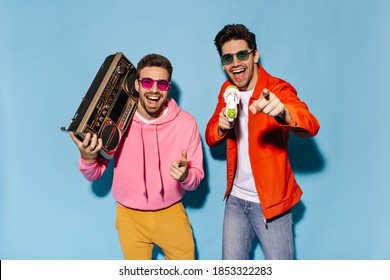 Joyful bearded friends in stylish outfits and sunglasses laugh and have fun on blue background. Guy in orange jacket holds water gun. Man in pink hoodie poses with record player.