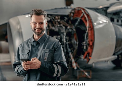 Joyful bearded man aircraft maintenance technician looking at camera and smiling while holding smartphone