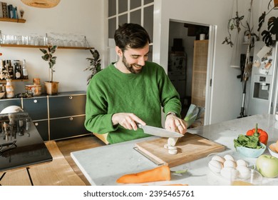 Joyful bearded man in green jumper cutting fresh leek on board at home - Shutterstock ID 2395465761