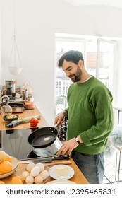 Joyful bearded man in green jumper holding frying pan with butter while cooking breakfast - Shutterstock ID 2395465831