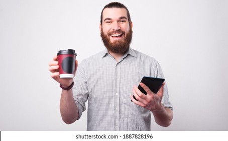A joyful bearded man is holding his coffe and a tablet having his coffee break smiles at the camera