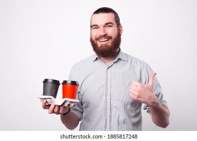 Joyful bearded man is smiling at the camera showing a thumb up because he likes the hot drink that he is holding in the papper cups near a white wall