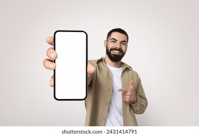 Joyful bearded millennial arab man in a casual khaki shirt and white t-shirt smiling and pointing at a smartphone with a blank white screen, suggesting app promotion or advertisement - Powered by Shutterstock