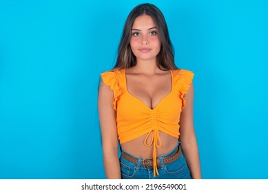 Joyful Beautiful Brunette Woman Wearing Orange Tank Top Over Blue Background Looking To The Camera, Thinking About Something. Both Arms Down, Neutral Facial Expression.