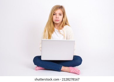 Joyful Beautiful Caucasian Teen Girl Sitting With Laptop In Lotus Position On White Background Looking To The Camera, Thinking About Something. Both Arms Down, Neutral Facial Expression.