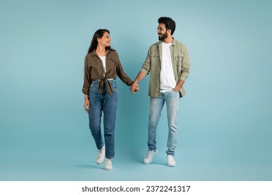 Joyful beautiful young eastern couple handsome bearded guy and long-haired pretty woman walking over plain blue background, holding hands, looking at each other and smiling, full length - Powered by Shutterstock