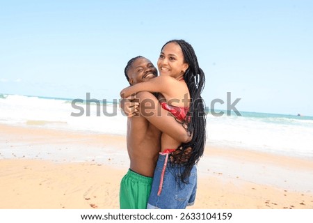 Joyful Black couple embracing at the beach. The woman is lifted off the ground, smiling brightly in a bikini top and denim shorts. The man, shirtless in green shorts, holds her with affection and laug