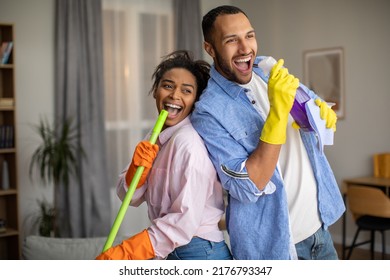 Joyful Black Couple Having Fun Cleaning House And Singing Favorite Song Holding Mop And Detergent Bottle Standing At Home. Family Lifestyle And Housework Concept