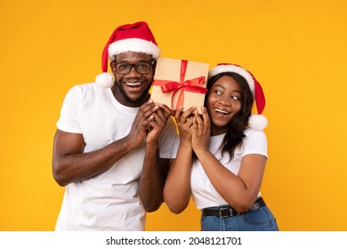 Joyful Black Couple Holding Christmas Gift Wrapped In Craft Paper, Listening To Box Guessing What's Inside Posing Over Yellow Background, Wearing Santa Hats. Studio Shot - Powered by Shutterstock