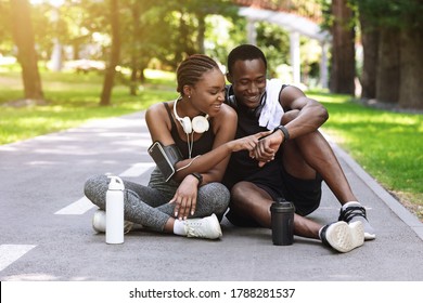 Joyful black couple resting after jogging in park, checking smartwatch fitness tracker while sitting on path, enjoying training outdoors, free space
