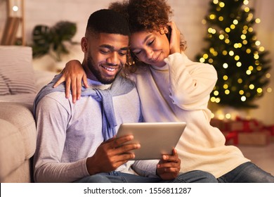 Joyful Black Couple Using Digital Tablet Against Christmas Tree, Enjoying Winter Evening At Home