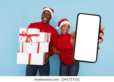 A joyful black couple wearing red sweaters and Santa hats poses with wrapped gifts, displaying a smartphone. They share festive energy and excitement for the holiday season. - Powered by Shutterstock