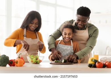 Joyful Black Family Making Fresh Vegetable Salad For Dinner Cutting Cucumbers In Modern Kitchen Indoor On Weekend, Wearing Aprons. Healthy Nutrition, Food Recipes Concept