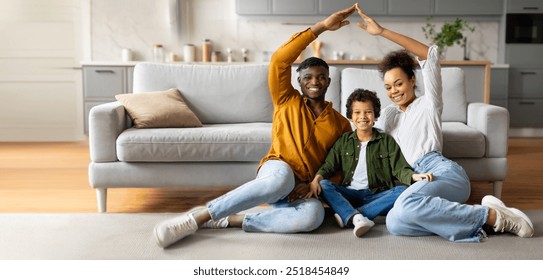 A joyful black family sits together on the floor, creating a playful atmosphere. The parents and child smile and make fun shapes with their hands, surrounded by a welcoming home environment. - Powered by Shutterstock