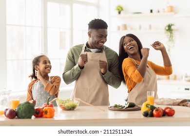 Joyful Black Family Of Three Dancing While Cooking, Having Fun Together Enjoying Dinner Preparation Standing In Kitchen Indoors. Happy Parents And Daughter Preparing Salad On Weekend