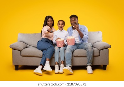 Joyful black family of three sitting on sofa and watching movie, eating popcorn from bucket laughing at show, woman holding remote control isolated on yellow studio wall - Powered by Shutterstock