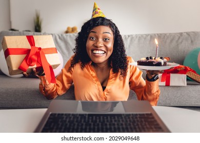 Joyful Black Female Celebrating Birthday Online Posing At Laptop With B-Day Cake And Wrapped Gift, Wearing Festive Hat And Smiling To Camera At Home. Remote Holiday Celebration