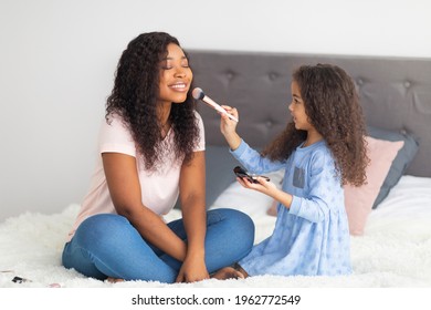 Joyful Black Girl Applying Makeup On Her Mom's Face On Bed At Home. African American Mother With Her Daughter Having Family Spa Day, Putting On Different Cosmetics, Enjoying Weekend Together