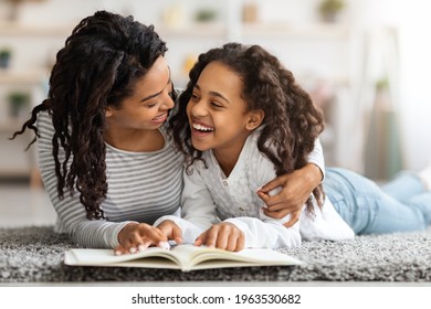 Joyful black girl reading book with her mom, laying together on floor and laughing, home interior. Happy african american mother and daughter reading books and embracing, closeup
