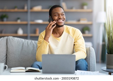 Joyful black guy freelancer having phone conversation while working from home, using laptop, smiling and looking at copy space. Young african american businessman talking with clients on mobile phone