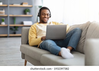 Joyful Black Guy Reclining On Couch At Home, Watching Movie On Laptop, Using Headset, Copy Space. Happy African American Man Playing Video Games Or Having Video Call While Resting On Weekend