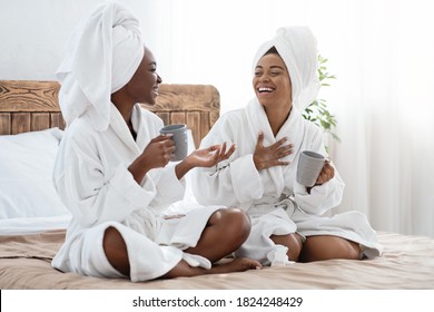 Joyful black ladies in bathrobes drinking coffee and laughing, having spa procedures at home. Two cheerful african american young women with cups of coffee having conversation in bedroom, empty space