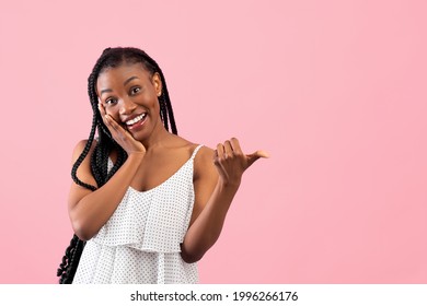 Joyful black lady pointing at blank space, touching her face in excitement over pink studio background. Charming African American woman advertising your product, promoting sale or discount