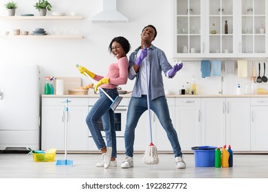 Joyful black loving couple singing songs while cleaning kitchen, using broom and mop as microphones and guitar, cheerful african american young man and woman imitating rock stars while house-keeping