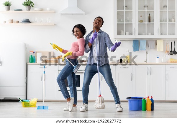 Joyful black loving couple singing songs\
while cleaning kitchen, using broom and mop as microphones and\
guitar, cheerful african american young man and woman imitating\
rock stars while\
house-keeping