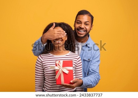 Joyful black man covering girfriends eyes for surprise and giving red gift box with white ribbon, happy young african american couple standing together against yellow studio background, copy space