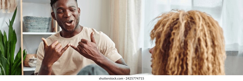 joyful black man smiling while communicating with sign language with girlfriend at home, banner