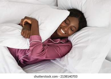 Joyful Black Millennial Lady Embracing Pillow Smiling With Eyes Closed Lying In Bed Wearing Pajamas Resting In Modern Bedroom At Home. Healthy Sleeping Routine, Comfort Concept