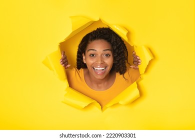 Joyful Black Millennial Woman Posing In Torn Paper Looking At Camera Through Hole Over Yellow Studio Background. Wow Offer Advertisement Concept. Collage