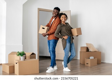 Joyful Black Spouses Holding Cardboard Moving Boxes Standing Back To Back Posing Smiling To Camera In New House Indoor. Real Estate Ownership, Family Housing Concept