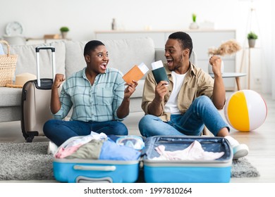 Joyful Black Tourists Couple Holding Tickets And Shaking Fists Celebrating Vacation Packing Suitcase At Home. Happy Tourists Preparing For Journey. Summer Tourism Concept. Selective Focus