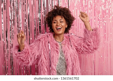 Joyful black woman dances actively, makes movements, raises hands, wears fur coat, smiles broadly, enjoys favourite music, has fun on shindig, gets together with friends. Celebration concept - Shutterstock ID 1361561702