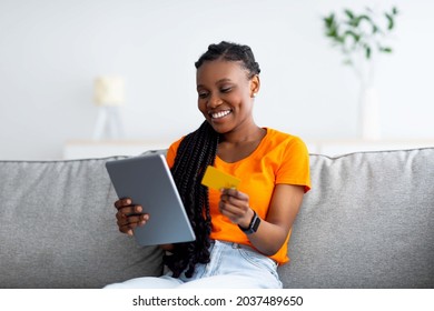 Joyful black woman making online payment, buying goods on web, using internet bank services, sitting on couch at home with tablet pc and credit card. Internet shopping, seasonal sale, black Friday