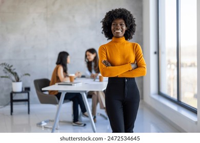 A joyful Black woman stands with arms crossed in a sunlit office, wearing professional attire, as diverse colleagues collaborate in the background. - Powered by Shutterstock