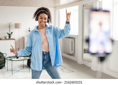 Joyful Black Woman In Wireless Headseat Dancing At Camera Filming Video Using Phone On Tripod At Home Creating Trendy Content On Mobile App To Share On Social Media, Selective Focus On Millennial Lady
