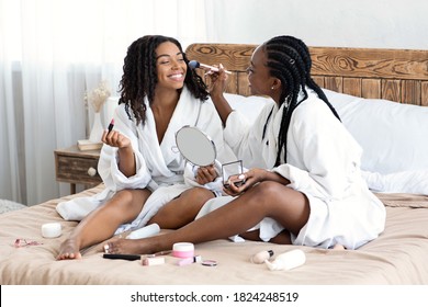 Joyful black women in bathrobes sitting on bed at home, putting makeup, looking at mirror. Attractive african american female friends having fun together at home, trying beauty products, copy space - Powered by Shutterstock
