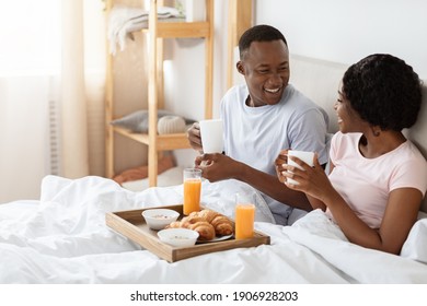 Joyful Black Young Couple Sitting In Bed In Morning And Having Breakfast, Drinking Coffee And Talking, Side View, Copy Space. Cheerful African American Man And Woman Enjoying Breakfast In Bed