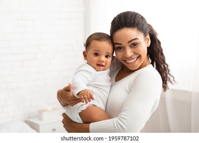 Joyful Black Young Mom Holding And Hugging Adorable Baby Boy Enjoying Motherhood And Child Care Standing In Modern Bedroom At Home, Smiling To Camera. Mother's Day Concept